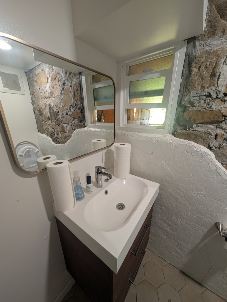 A modern bathroom view featuring a sink with a faucet, a wall mirror, and two rolls of paper towels. Stone wall texture is visible behind the sink, and natural light comes through small windows.