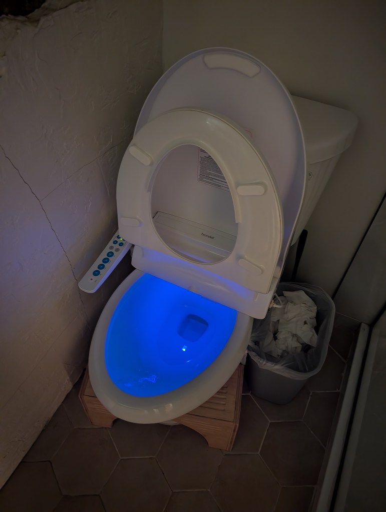 A modern toilet with a glowing blue interior, featuring a remote control on the side and a waste bin nearby on tiled flooring.