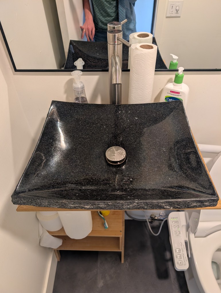 A modern black granite sink with a sleek faucet, accompanied by paper towels, soap dispensers, and a mirror.