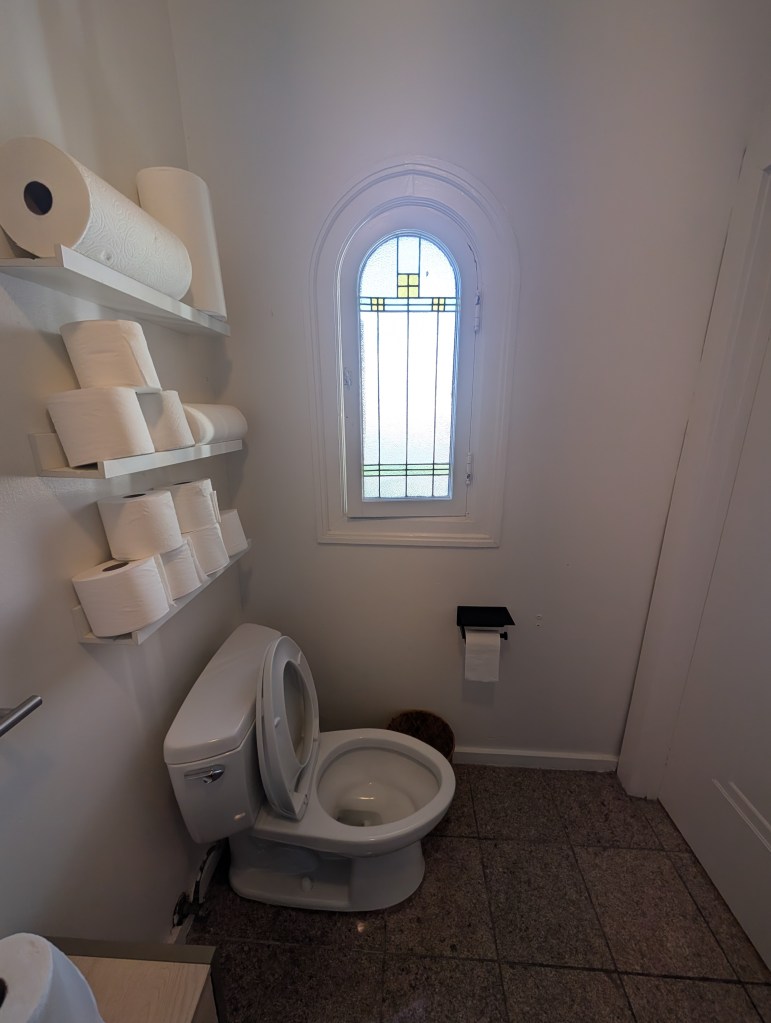 A small bathroom featuring a modern toilet, a stained glass window, and shelves stocked with toilet paper rolls.